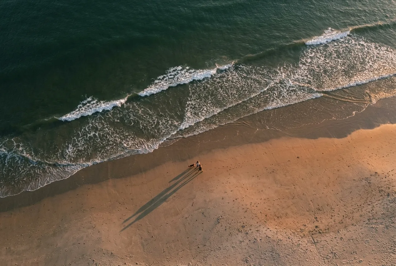 Luchtfoto van golven op een breed strand met een wandelend stel en hond