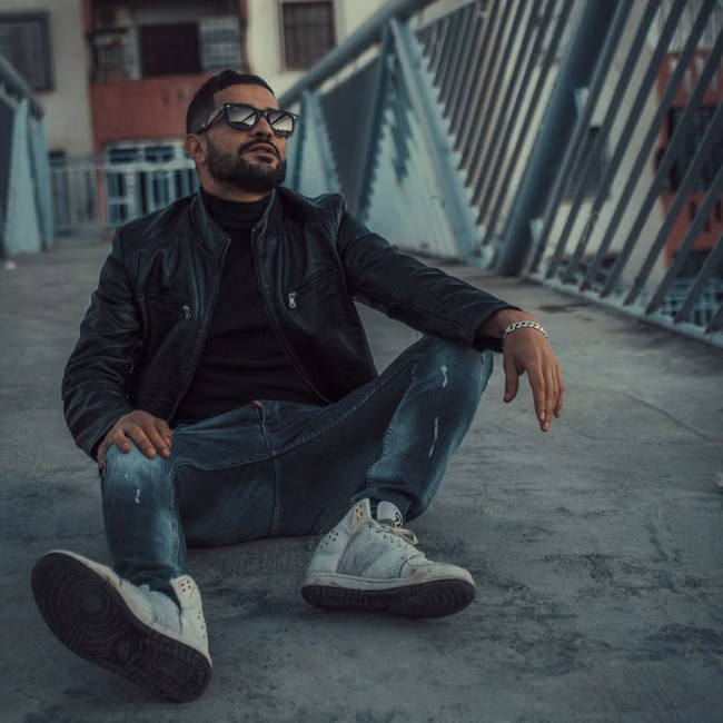 Fashionable man in leather jacket and sunglasses sitting on a bridge, exuding casual style.